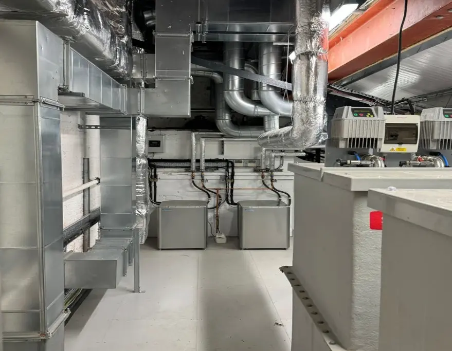 Basement plant room with galvanised and insulated ductwork overhead, water heaters against the back wall, booster pump controls and cold water storage tanks