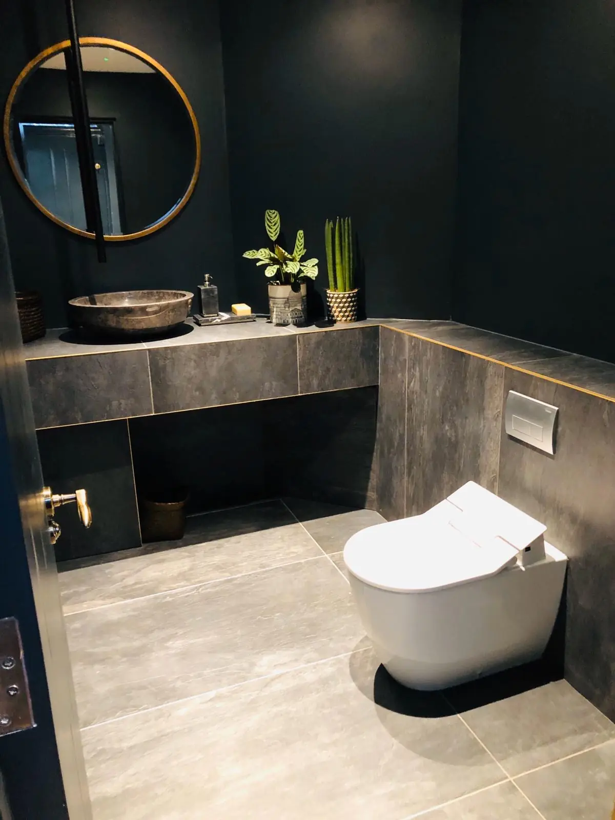 Cifonelli Mayfair WC with dark walls, slate-effect tiling, stone vessel basin, round brass mirror and shower toilet