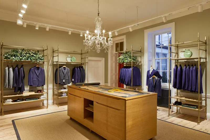 Hackett Savile Row bespoke room with oak cutting table, brass rails of navy suits, chandelier and column radiator